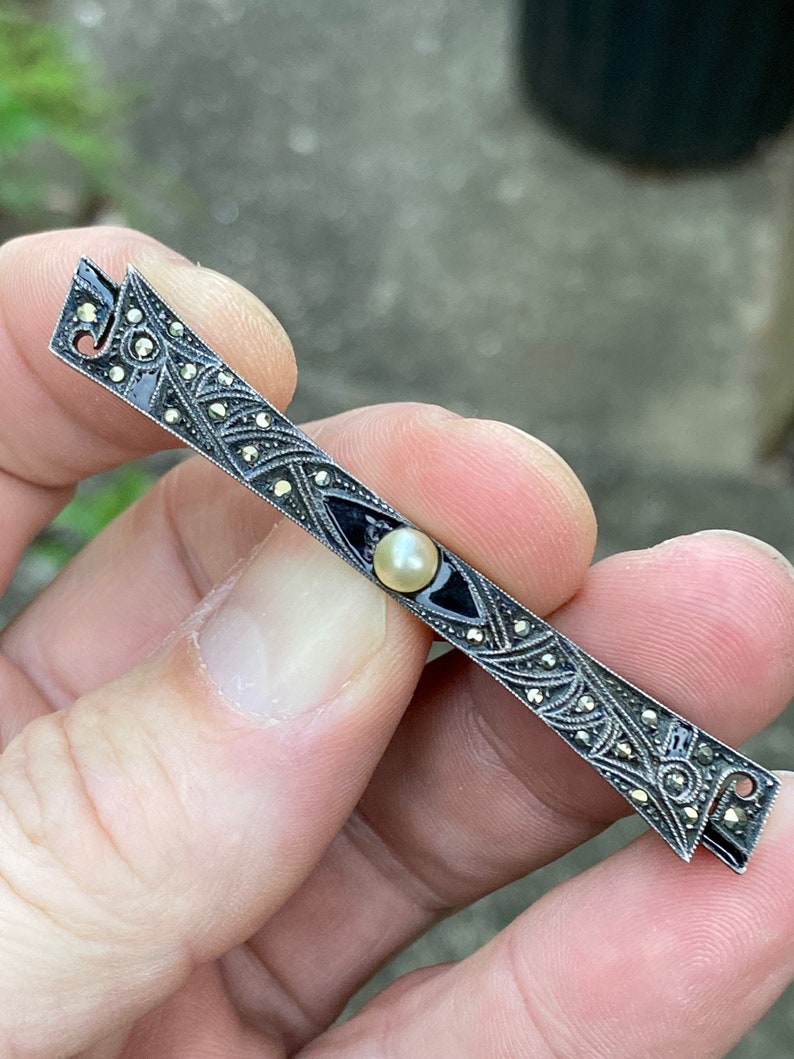 May include: A silver bar pin with a black enamel center and a single pearl. The pin is decorated with marcasite stones.