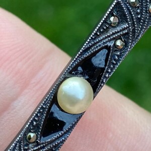 May include: A silver and black brooch with a pearl in the center. The brooch is decorated with a geometric pattern and small black stones.