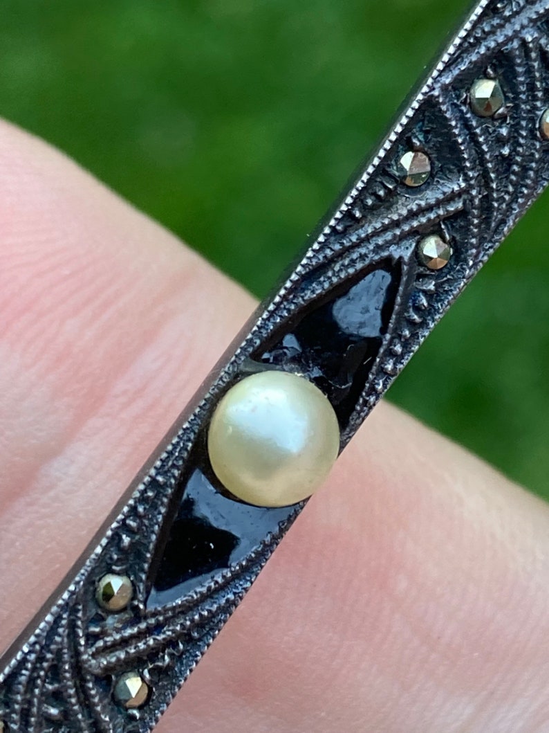 May include: A silver and black brooch with a pearl in the center. The brooch is decorated with a geometric pattern and small black stones.