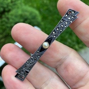 May include: A silver bar pin with black enamel and a pearl accent. The pin is decorated with a geometric design and is set with marcasite stones.