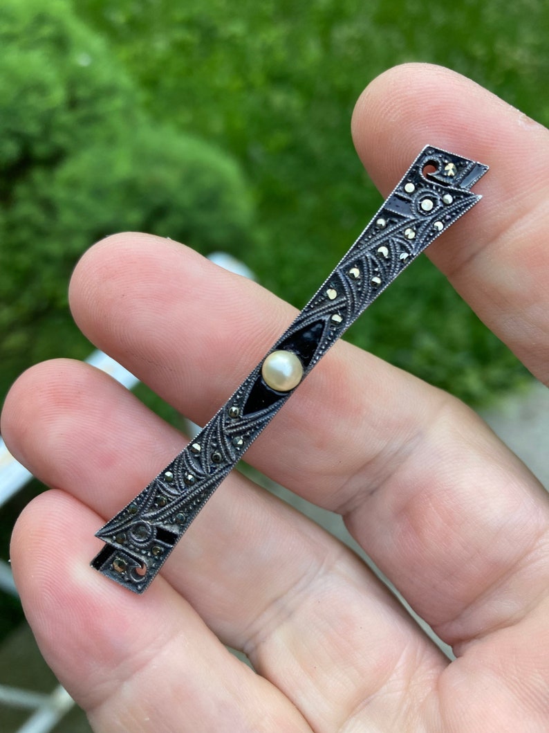 May include: A silver bar pin with black enamel and a pearl accent. The pin is decorated with a geometric design and is set with marcasite stones.