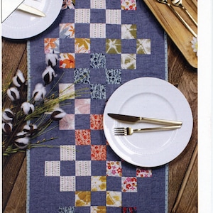 May include: A patchwork table runner with a blue background and colorful squares. The runner is laid out on a wooden table with two place settings, each with a white plate, gold silverware, and a cotton boll arrangement.