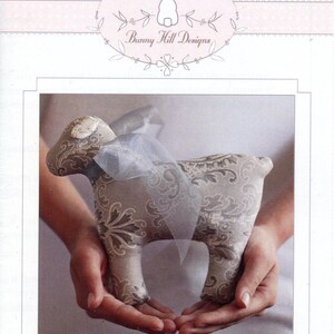May include: A stuffed lamb toy with a damask pattern in shades of beige and brown, held in open hands. The lamb has a light blue ribbon tied around its neck. The image includes the text "Bunny Hill Designs" and "Lollipop".