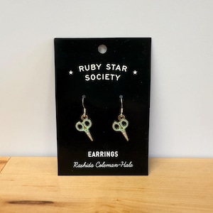 May include: A pair of gold-colored earrings shaped like scissors, with mint green and white accents. The earrings are displayed on a black card with the text "RUBY STAR SOCIETY" and "EARRINGS" and the name "Rashida Coleman-Hale".