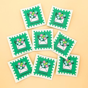May include: A collection of square fabric labels with a green postage stamp design. Each label features a white envelope overflowing with colorful flowers and the word "HANDMADE" in white text.