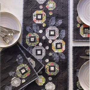 May include: A quilting pattern for a table runner and four placemats. The pattern features a black background with a geometric design of white and gray squares and circles. The design is called "Posies" and is by Robin Pickens Quilt Patterns.