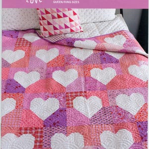 May include: A patchwork quilt with large white heart shapes on a pink, purple, and red background. The quilt is made of various patterned fabrics. The image also shows a matching pillow with a geometric design. The text on the image says "Patchwork Hearts II".
