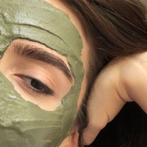 May include: A close-up of a person's face with a green clay face mask applied. The mask is covering most of the face, leaving only the eyes and eyebrows visible. The person's hair is brown and is partially covering the face.