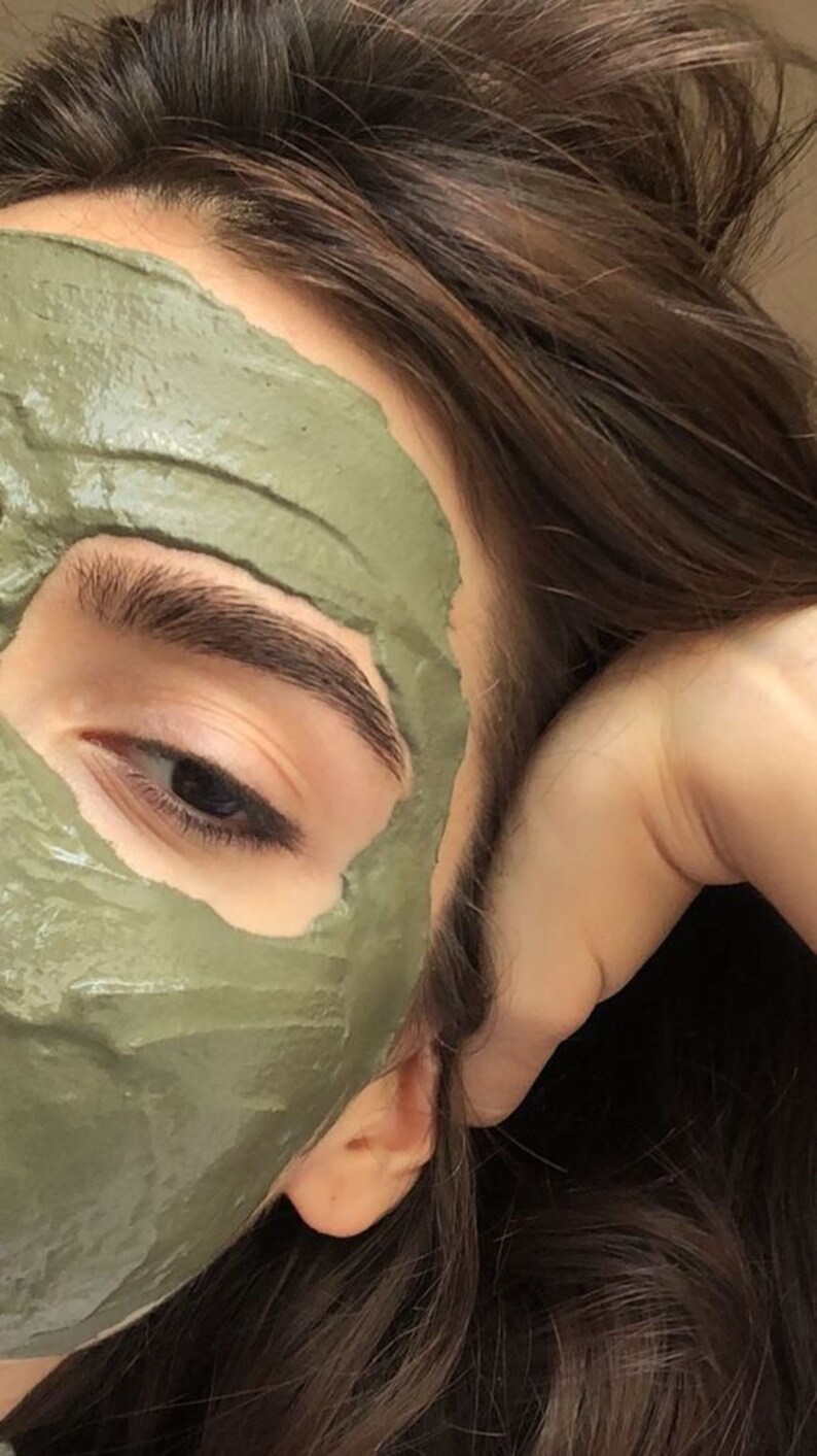 May include: A close-up of a person's face with a green clay face mask applied. The mask is covering most of the face, leaving only the eyes and eyebrows visible. The person's hair is brown and is partially covering the face.