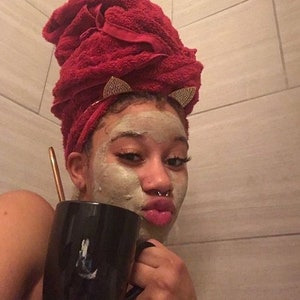 May include: A person with a face mask on is holding a black mug with a white design. They are wearing a red towel on their head with gold cat ear clips.