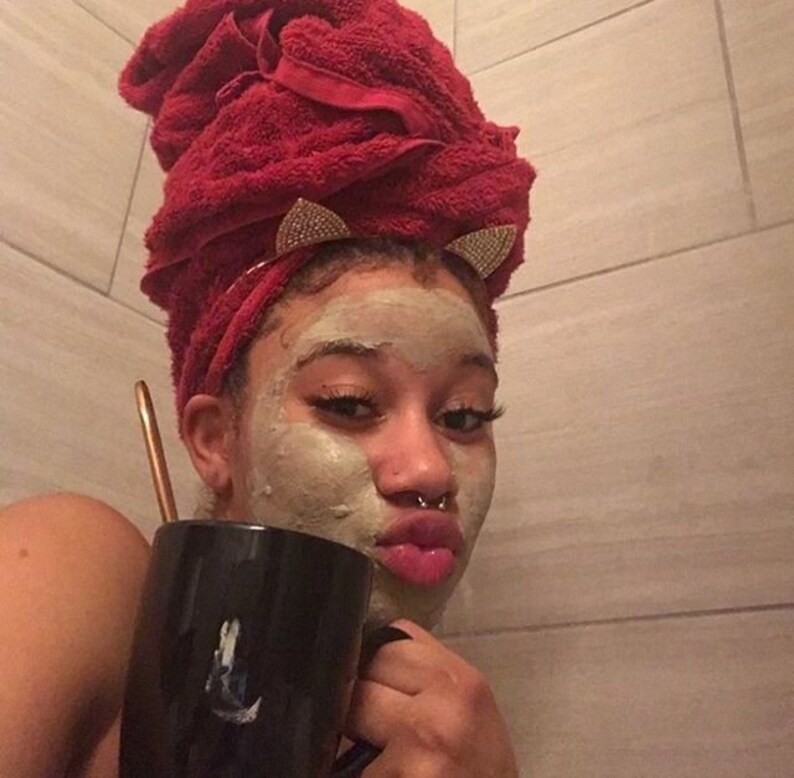 May include: A person with a face mask on is holding a black mug with a white design. They are wearing a red towel on their head with gold cat ear clips.
