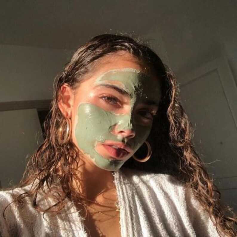 May include: A woman with long, curly brown hair is wearing a green face mask. She is wearing a white robe and has a gold necklace on.
