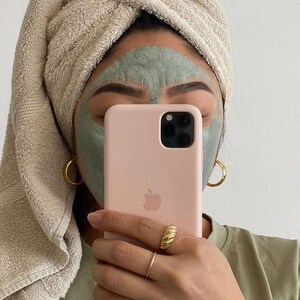 May include: A person with a green face mask holds a pink iPhone with a white Apple logo. The phone case has a camera lens cutout. The person is wearing a beige towel wrapped around their head and a green shirt.