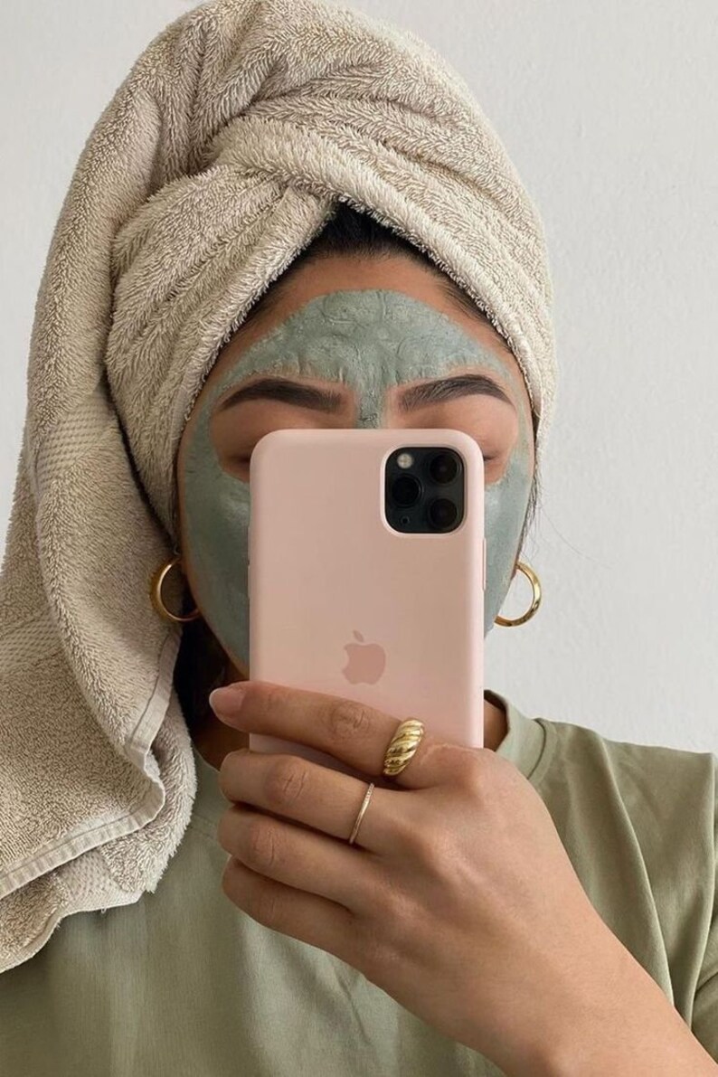 May include: A person with a green face mask holds a pink iPhone with a white Apple logo. The phone case has a camera lens cutout. The person is wearing a beige towel wrapped around their head and a green shirt.
