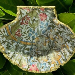 Può includere: Conchiglia decorativa a forma di capasanta con un motivo floreale nei toni del verde, blu, rosso e giallo. Il bordo della conchiglia è rifinito con vernice dorata, creando un pezzo unico. Appoggiata su un letto di foglie verdi.