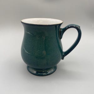 Puede incluir: Una taza de cerámica verde con interior blanco y borde oscuro. La taza tiene un cuerpo redondeado, una base de pedestal y un asa curva. La taza mide aproximadamente 10 cm de alto.