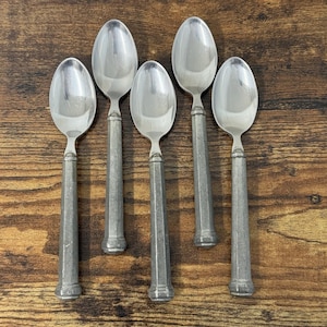 May include: A set of five silver-toned spoons arranged in a vertical row. Each spoon features a polished, reflective oval bowl and a long, matte-finished, cylindrical handle with a slightly flared base, creating a uniform appearance.