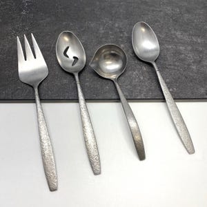 May include: A collection of four vintage stainless steel serving utensils. The set includes a fork, slotted spoon, ladle, and spoon. Each piece has a textured handle and a silver finish. Suitable for serving food.
