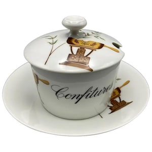 May include: White ceramic jam jar with lid and saucer. The jar features a brown and gold illustration of a cooking appliance and the word "Confitures" in cursive. The lid has a small knob and a spoon rest.