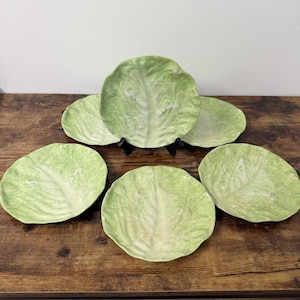 May include: A set of five ceramic plates shaped like cabbage leaves, featuring a light green color with realistic embossed vein textures. The plates have a scalloped edge and a glossy finish, arranged in a cluster to display their leaf-like design.