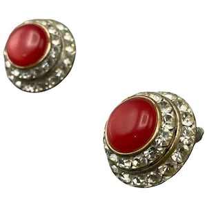 May include: Two vintage stud earrings. Each earring features a vibrant red, oval-shaped center stone surrounded by two concentric rings of clear, sparkling rhinestones. The earrings have a gold-tone metal setting.
