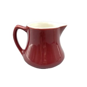 May include: A small, burgundy-colored ceramic creamer with a white interior. The creamer has a curved body, a handle, and a spout for easy pouring. The design is simple and classic, suitable for serving cream or milk.