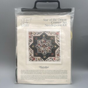 May include: A needlepoint kit for a coaster set, titled "Star of the Orient." The design features a central star shape with floral patterns in red, cream, and green, set against a dark background. The kit is packaged in a clear plastic bag.