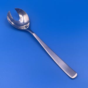 May include: A shiny, silver salad serving spoon with a unique, curved design. The spoon features a long, slender handle and a wide, slotted head, perfect for serving salads. The spoon is made of a reflective metal and is set against a blue background.