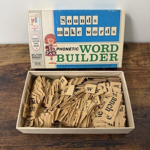 May include: A vintage Milton Bradley Phonetic Word Builder game box with a blue and cream cover featuring the text Sounds make words and a cartoon girl. The open box contains a large collection of small, tan cardboard tiles printed with black letters.