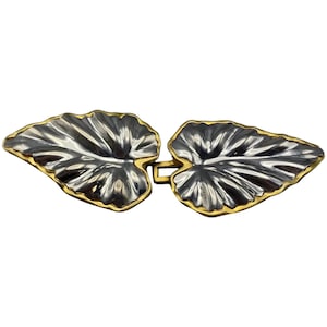 May include: A silver and gold-toned leaf-shaped clasp. The clasp is made of two stylized leaves with silver-coloured veins and a gold-coloured border. The leaves are connected by a gold-coloured rectangular clasp.