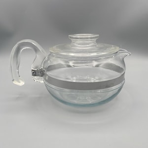 May include: A clear glass teapot with a rounded body, a curved handle, and a lid. A silver-coloured band encircles the middle of the teapot. The spout is on the right side. The teapot is designed for brewing tea.