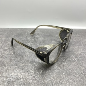 May include: Translucent grey-tinted glasses featuring round lenses and integrated black mesh side shields. The frames are constructed from a smooth, semi-transparent material with thin temples that extend from the side-mounted protective mesh panels.