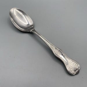 May include: A silver-toned metal spoon featuring a polished oval bowl and a handle decorated with an embossed shell-like motif at the tip and intricate linear detailing extending along the stem toward the neck of the spoon.
