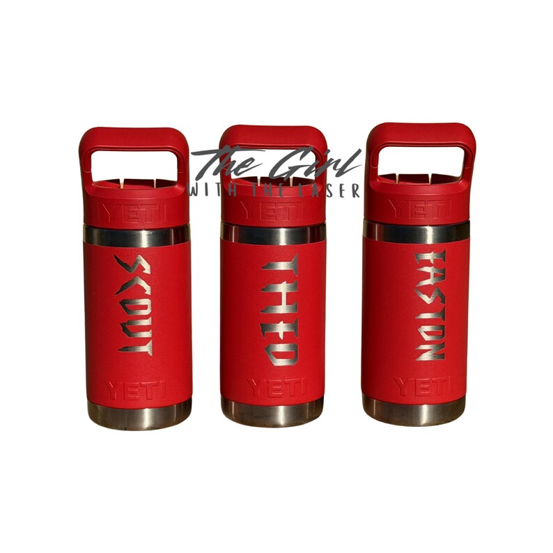 May include: Three red insulated tumblers with silver lettering. Each tumbler has a handle and the names "SCOUT", "THEO", and "TASTON" etched on the front. The brand name "YETI" is visible on each tumbler.