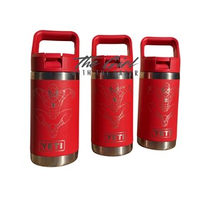 May include: Three red insulated water bottles with silver accents and red handles. Each bottle features a silver Spider-Man graphic and the brand name "YETI". The text "The Girl Within The Air" is visible.