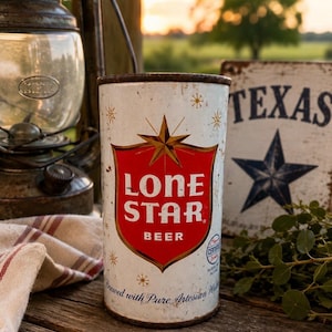 May include: A weathered Lone Star Beer can featuring a red and gold star emblem on a white base. The can, showing signs of age, is placed on a wooden surface. A 'Texas' sign is visible in the background. The can's text reads "Brewed with Pure Artesian Water."