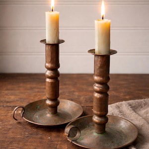 May include: Two antique copper candle holders with lit ivory candles. The candle holders have a hammered texture and a round base with a handle. The candles are cylindrical and have a warm glow, creating a cozy ambiance.