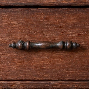 Puede incluir: Primer plano de un tirador de cajón de estilo antiguo, marrón oscuro. El tirador es de metal y tiene un diseño detallado y ornamentado. El tirador está montado en un cajón de madera con un rico acabado marrón rojizo. La veta de la madera es visible.