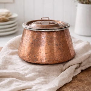 Puede incluir: Una olla de cobre con tapa, con una textura martillada. La olla es redonda con una base ligeramente ensanchada y un pequeño asa en la tapa. Descansa sobre un paño de lino beige, lo que sugiere una estética rústica o vintage. La olla tiene unos 15 cm de diámetro.