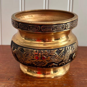 May include: A small, round, gold-colored metal container with intricate black dragon and geometric designs. The container has a wide opening and a decorative rim. Red accents are visible on the dragon design. The container sits on a wooden surface.