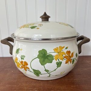 Op de afbeelding: Een witte vintage emaille pot met deksel, versierd met gele en oranje bloemen en groene bladeren. Het heeft houten handvatten en een houten knop. De rand is zilverkleurig. Ongeveer 25 cm in diameter.
