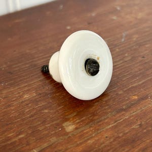 Pode incluir: Um puxador de armário de cerâmica branca com uma face redonda e plana e uma base menor e arredondada. O puxador tem um parafuso preto e está colocado sobre uma superfície de madeira. O puxador é provavelmente para uma gaveta ou armário.