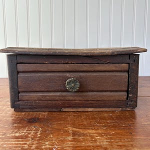 Könnte beinhalten: Kleine, antike Holzschublade mit dunkelbraunem Finish. Die Schublade hat einen dekorativen Metallknopf und steht auf einer Holzoberfläche. Das Holz weist Alters- und Gebrauchsspuren auf, was ihren Vintage-Charme unterstreicht. Die Schublade dient wahrscheinlich zur Aufbewahrung.