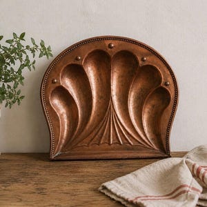 May include: A copper-colored, decorative wall hanging with a stylized, symmetrical design. The piece has a rounded top and features a series of curved, petal-like shapes. Small, round rivets and a textured surface add detail. The item is displayed on a wooden surface.