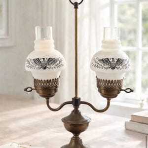 May include: A vintage-style table lamp with two frosted glass shades, each adorned with a black eagle emblem and floral accents. The lamp features a bronze-colored metal base and a central support with a top loop. The lamp is on a light-colored surface.