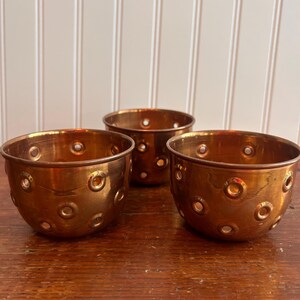 Peut inclure: Trois bols en métal de couleur cuivre avec une série de perforations circulaires espacées. Les bols sont disposés sur une surface en bois, avec un mur à panneaux blancs en arrière-plan. Les bols ont une forme arrondie.