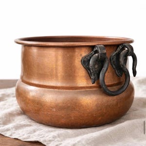 May include: A copper-colored metal pot with a rounded base and a wide rim. The pot features two ornate, dark-colored handles. The pot is sitting on a light-colored, textured fabric.