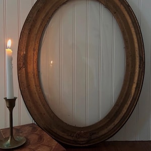 Può includere: Un'antica cornice ovale con finitura dorata, con dettagli decorati in alto e in basso. Una candela bianca accesa è posta in un candelabro di ottone a sinistra della cornice. Un libro vintage è sotto la cornice.
