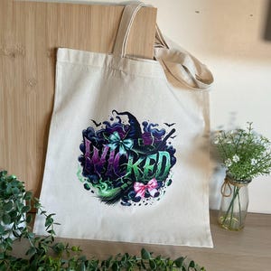 May include: A cream-coloured canvas tote bag featuring a graphic print of a witch hat, a broom, and the word WICKED in stylised purple and green lettering, accented with a light blue bow and a pink bow against a dark swirling background.