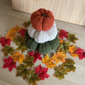 May include: A stack of three crocheted pumpkins in orange, white, and olive green, topped with a small green stem. The pumpkins are surrounded by a scattering of artificial autumn leaves in shades of red, yellow, orange, and green on a light surface.
